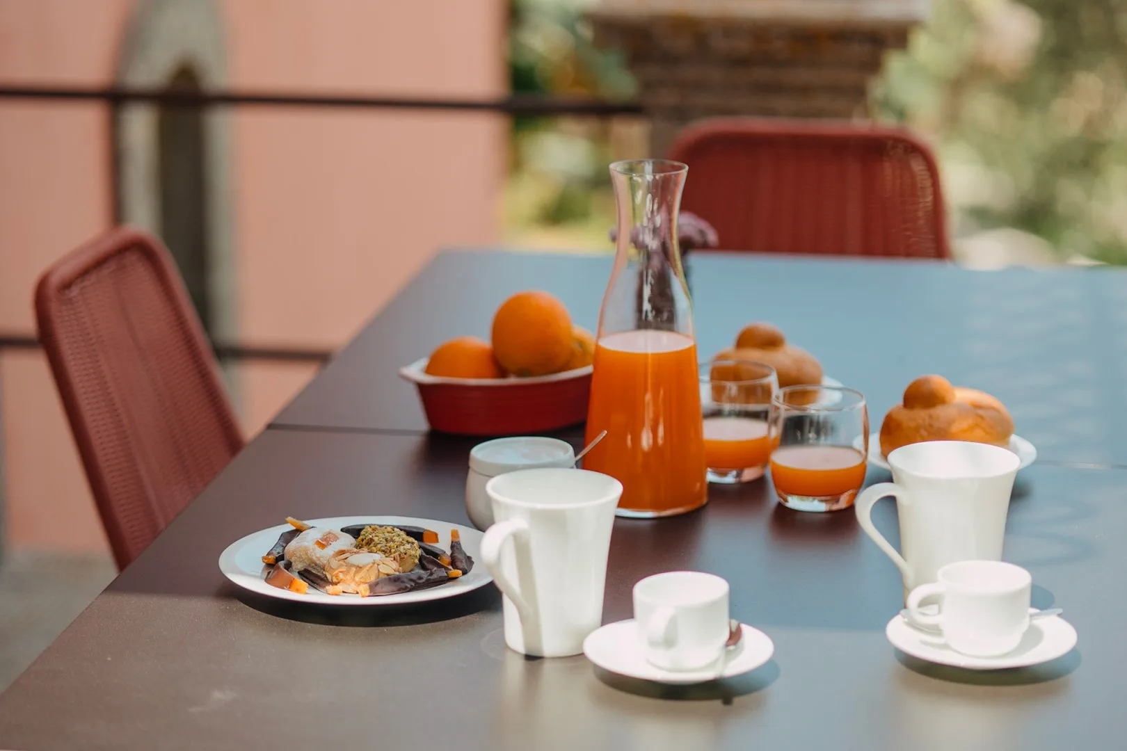Una mesa de comedor al aire libre está preparada con desayuno, zumo de naranja y tazas.