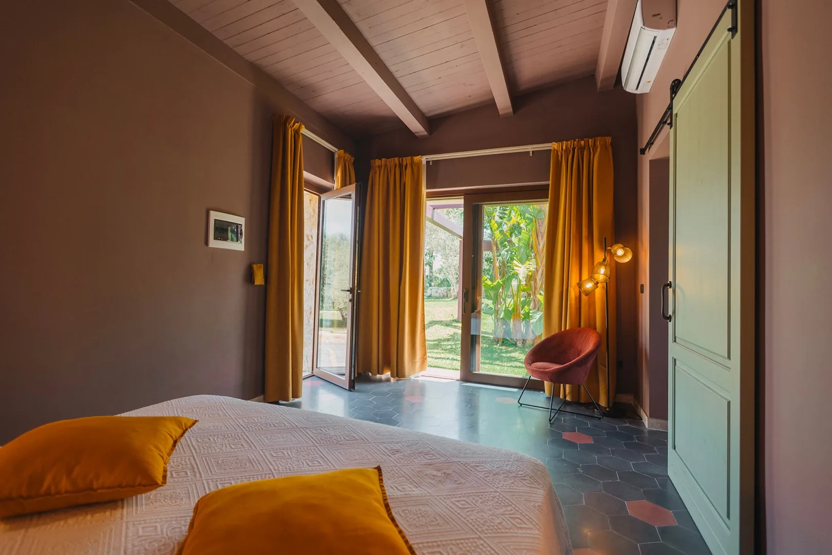 Un dormitorio con una cama, cortinas mostaza y puertas de vidrio que dan a la vegetación.
