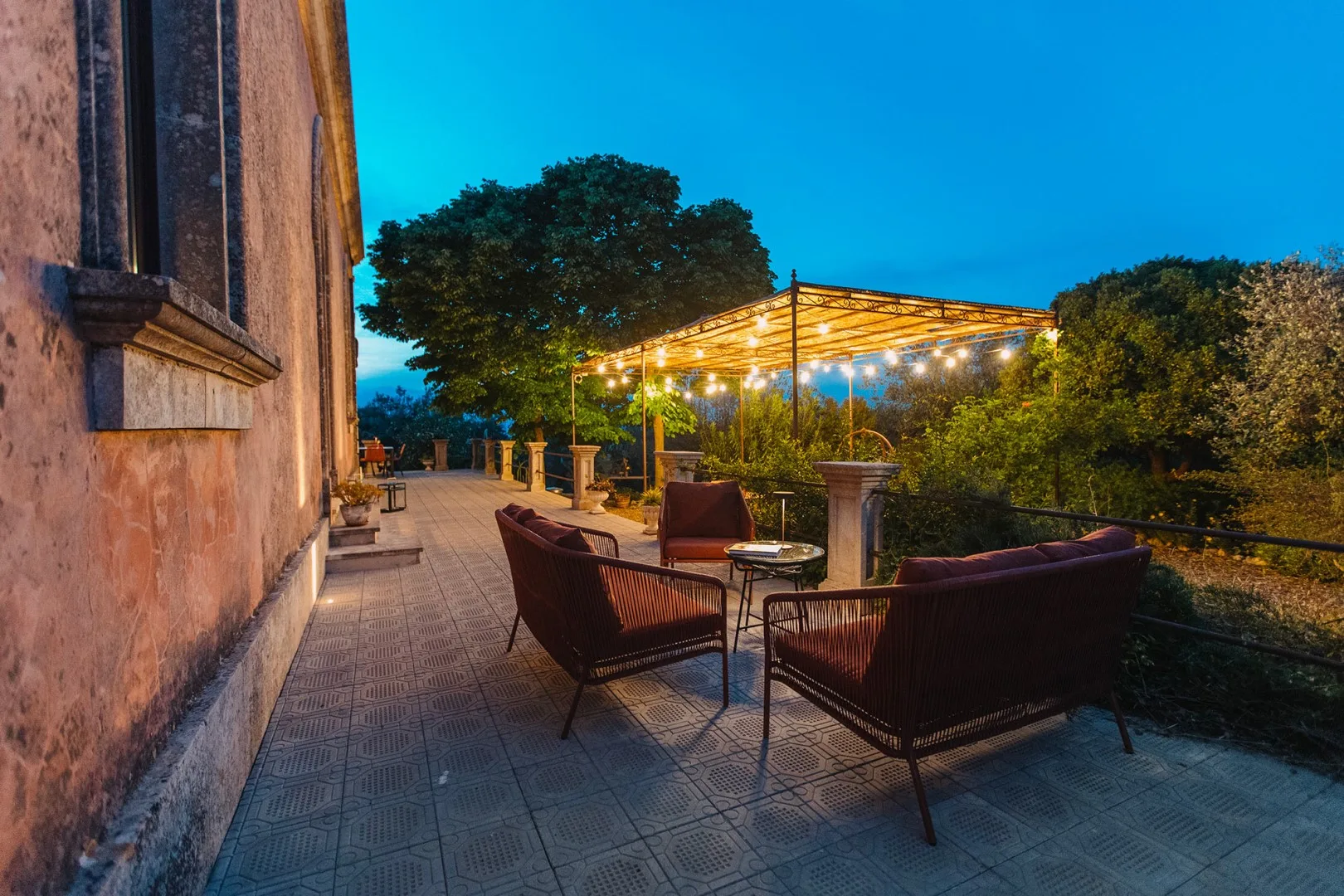 Un patio exterior tiene asientos bajo una pérgola con luces colgantes.