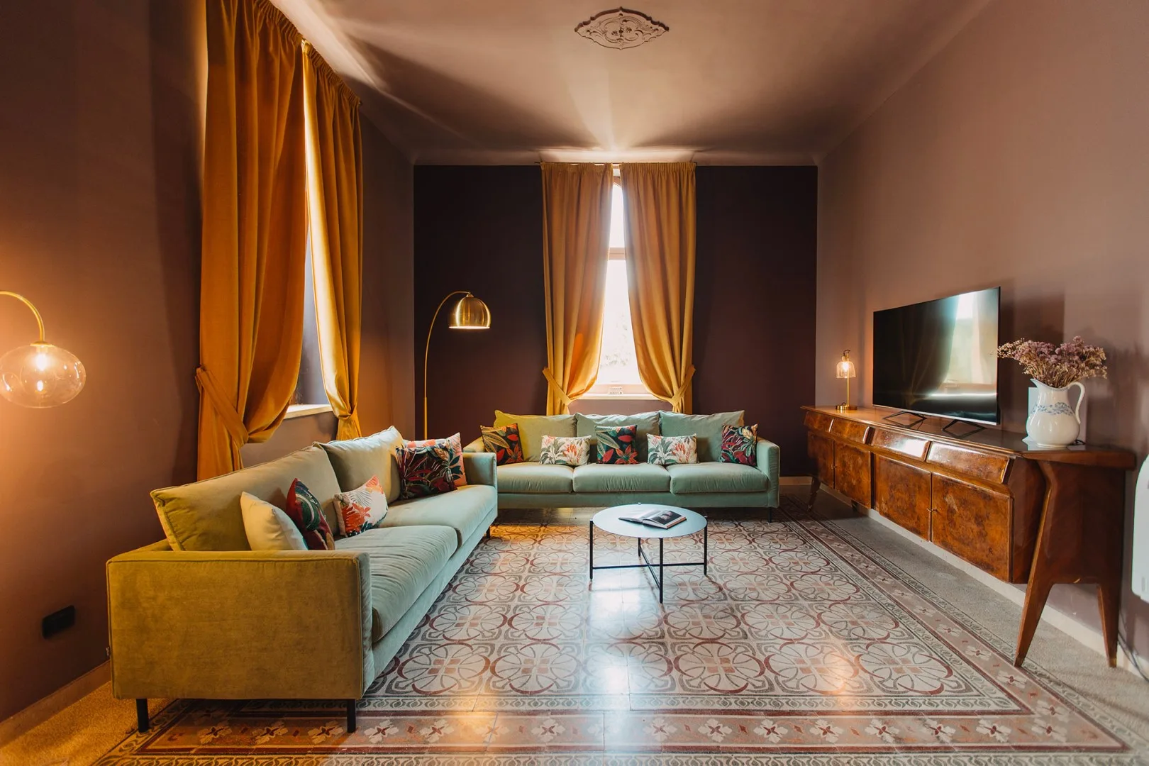 Una sala de estar con asientos verdes, cortinas mostaza y un televisor sobre una consola de madera.