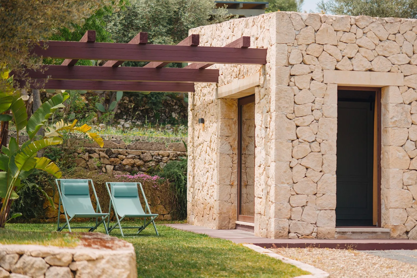 Una construcción de piedra está junto a una pérgola de madera y asientos en el jardín.