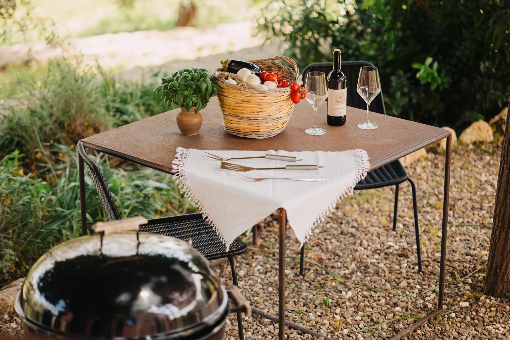 Una mesa de comedor al aire libre está puesta con vino, copas y una cesta de productos.