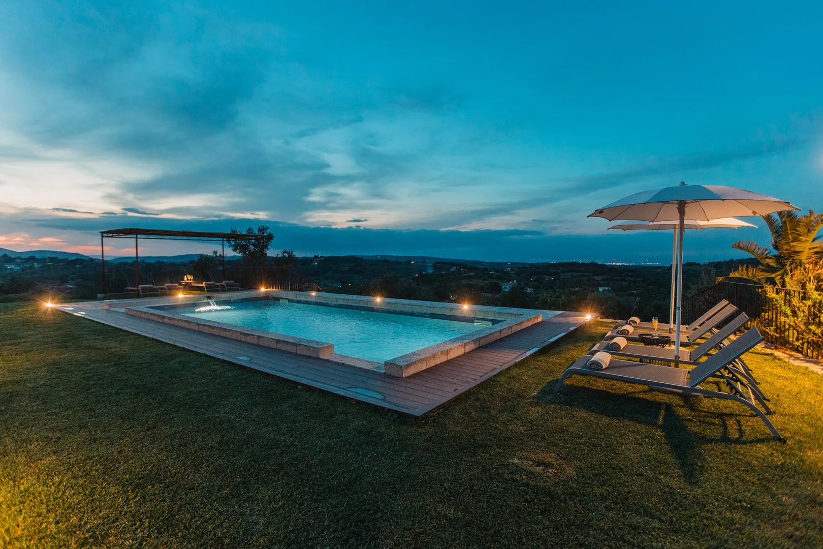 Una piscina al aire libre junto a asientos reclinables y una sombrilla abierta al atardecer.