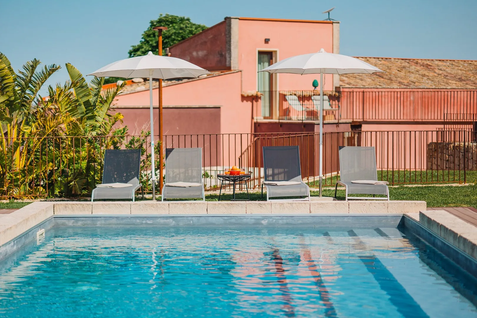 Una piscina está frente a un edificio rosa con tumbonas a la sombra.