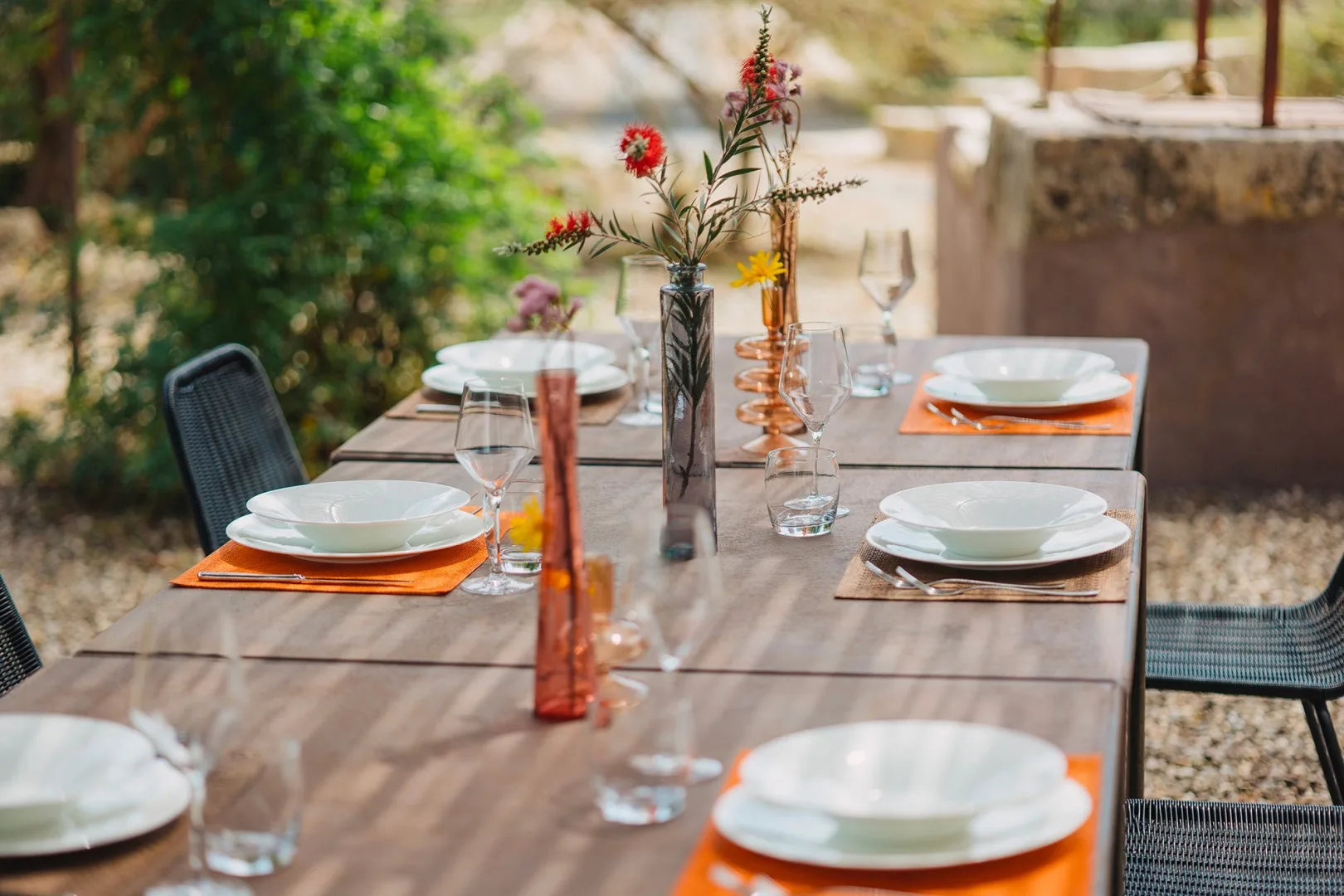 Una mesa de comedor al aire libre está puesta con platos, vasos y pequeños arreglos florales.