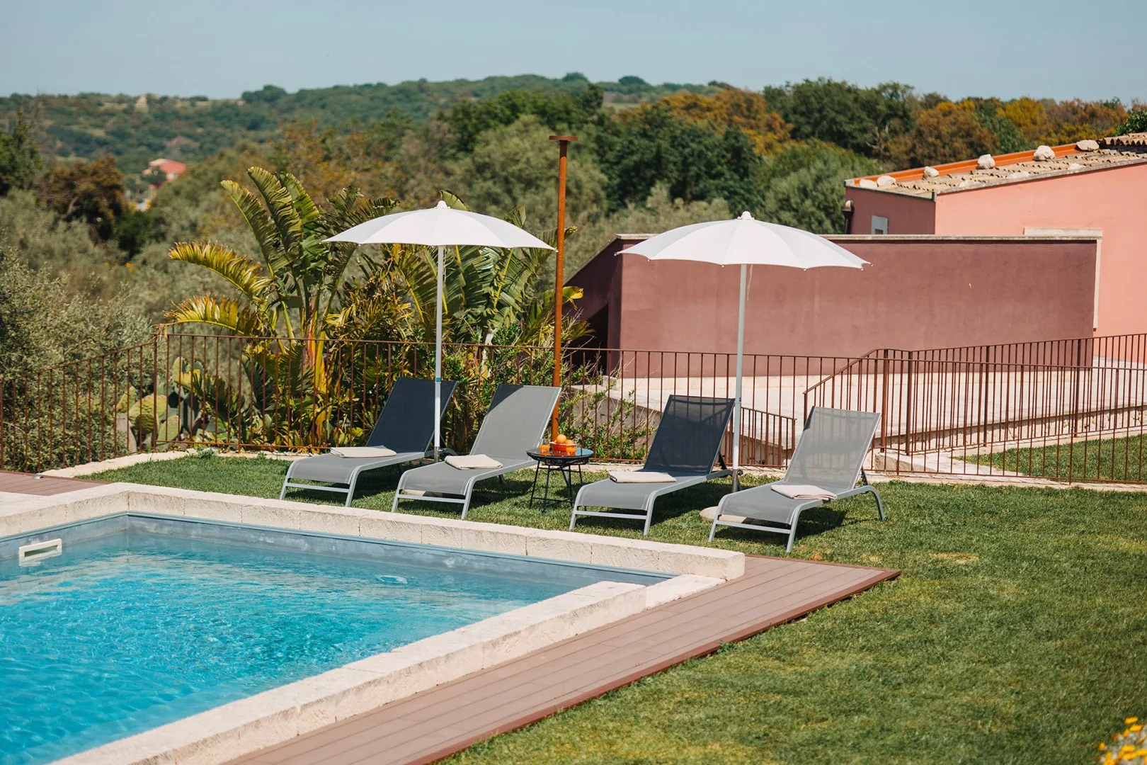 Una piscina al aire libre junto a asientos reclinables y sombrillas, con colinas arboladas al fondo.