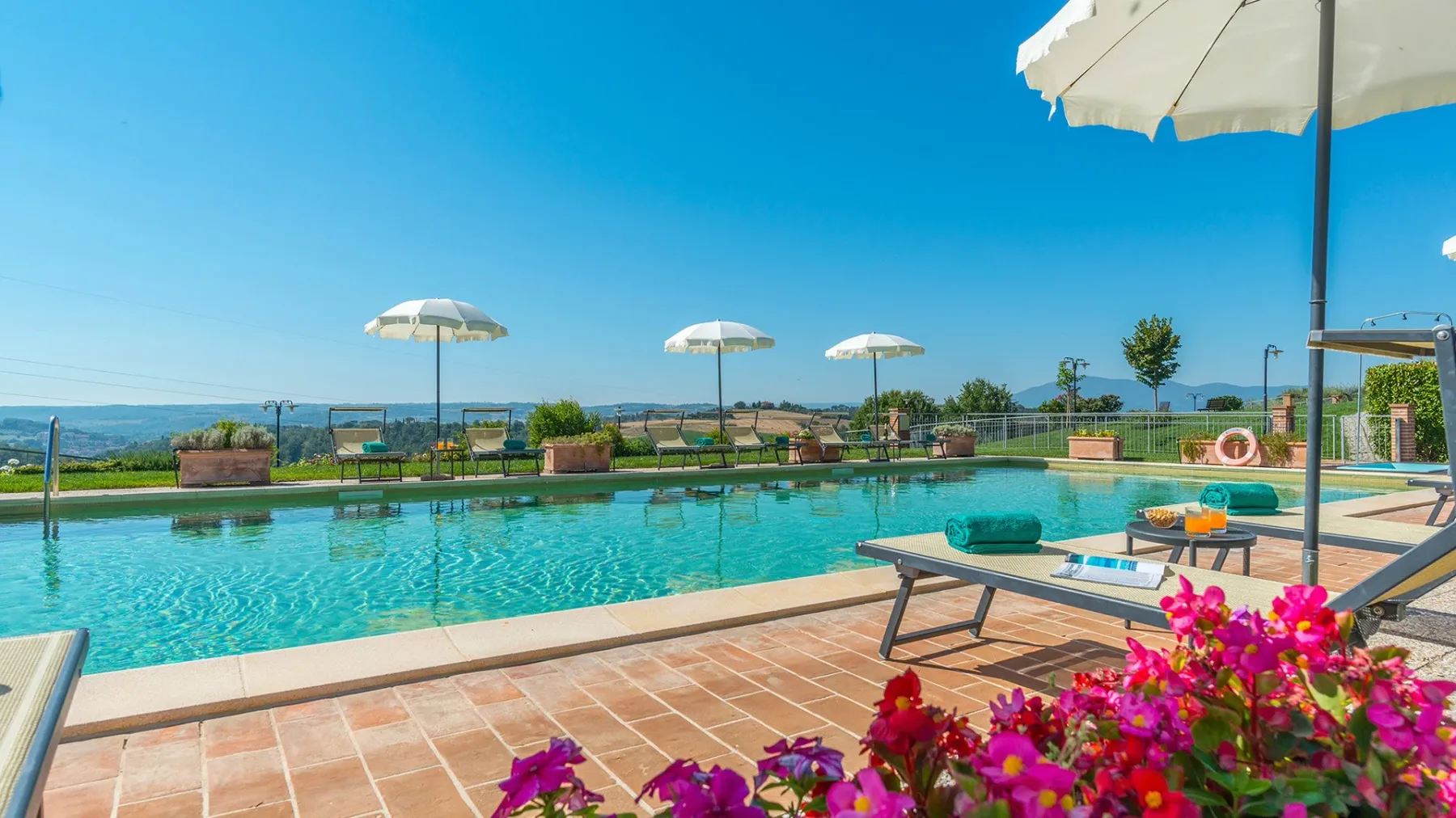 An outdoor swimming pool overlooks a green landscape, with lounge seating on the tiled deck.