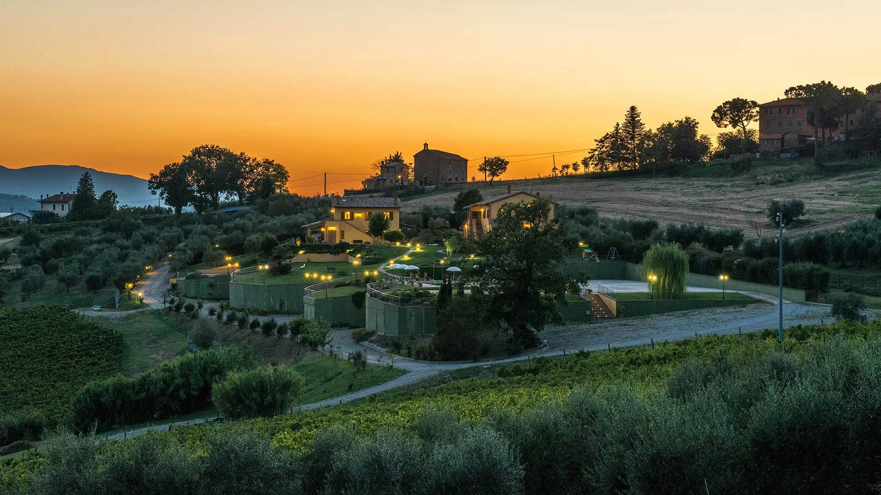 A rural hillside property sits among fields and trees at sunset.