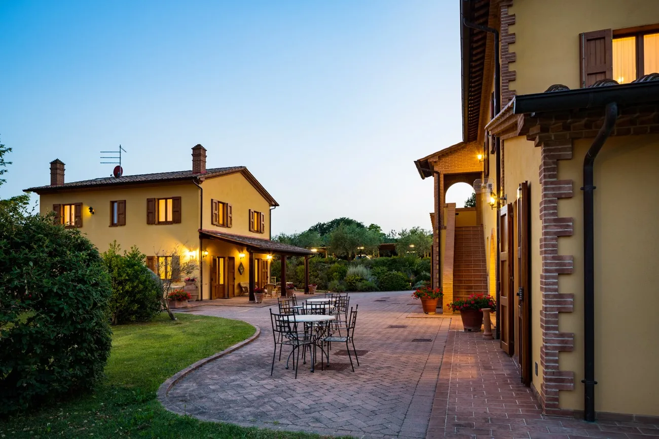 A brick patio with outdoor seating sits beside warmly lit buildings at dusk.