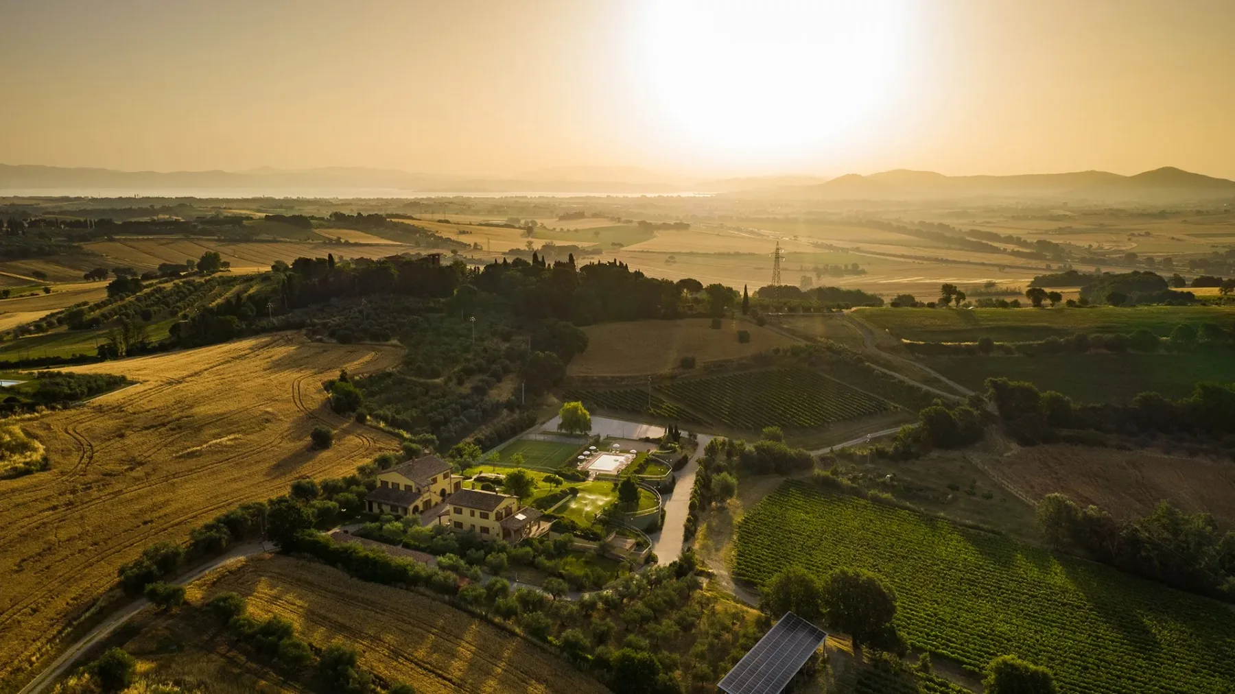 A rural landscape at sunset, with fields and a small cluster of buildings.