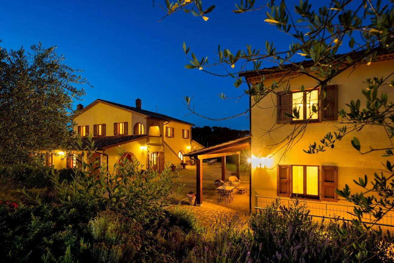 Two warmly lit houses stand beside a garden at dusk.
