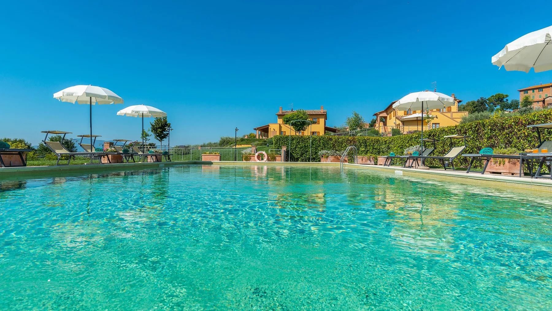 A swimming pool surrounded by sun loungers and umbrellas under a clear sky.