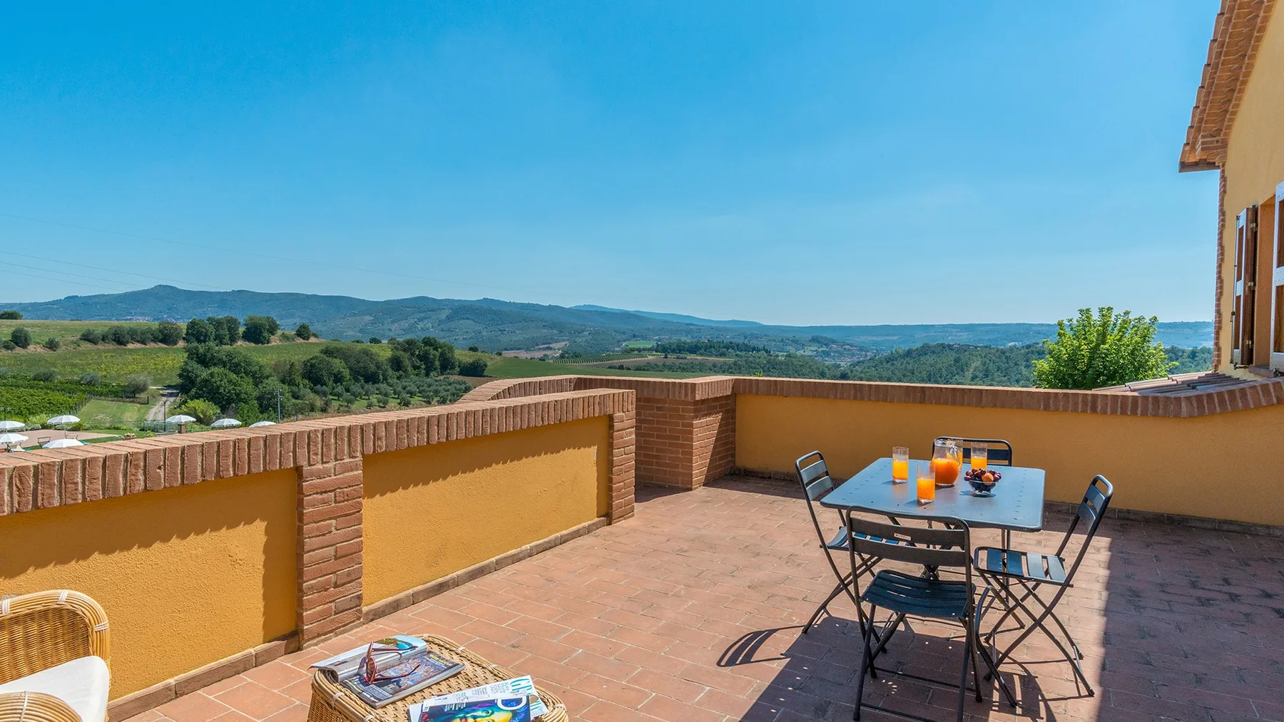 A tiled rooftop terrace has a dining table overlooking green fields and distant hills.
