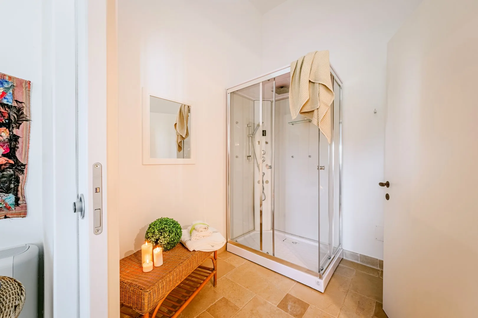 Un baño luminoso cuenta con una ducha esquinera de vidrio, un espejo de pared y un banco de mimbre con velas y una planta.
