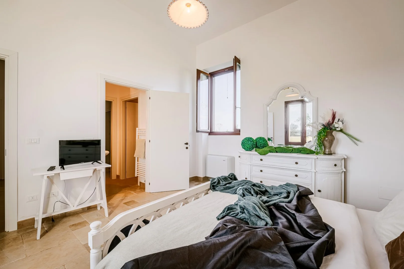 Un dormitorio luminoso tiene una cama blanca con ropa de cama oscura y arrugada, una cómoda y una ventana abierta.