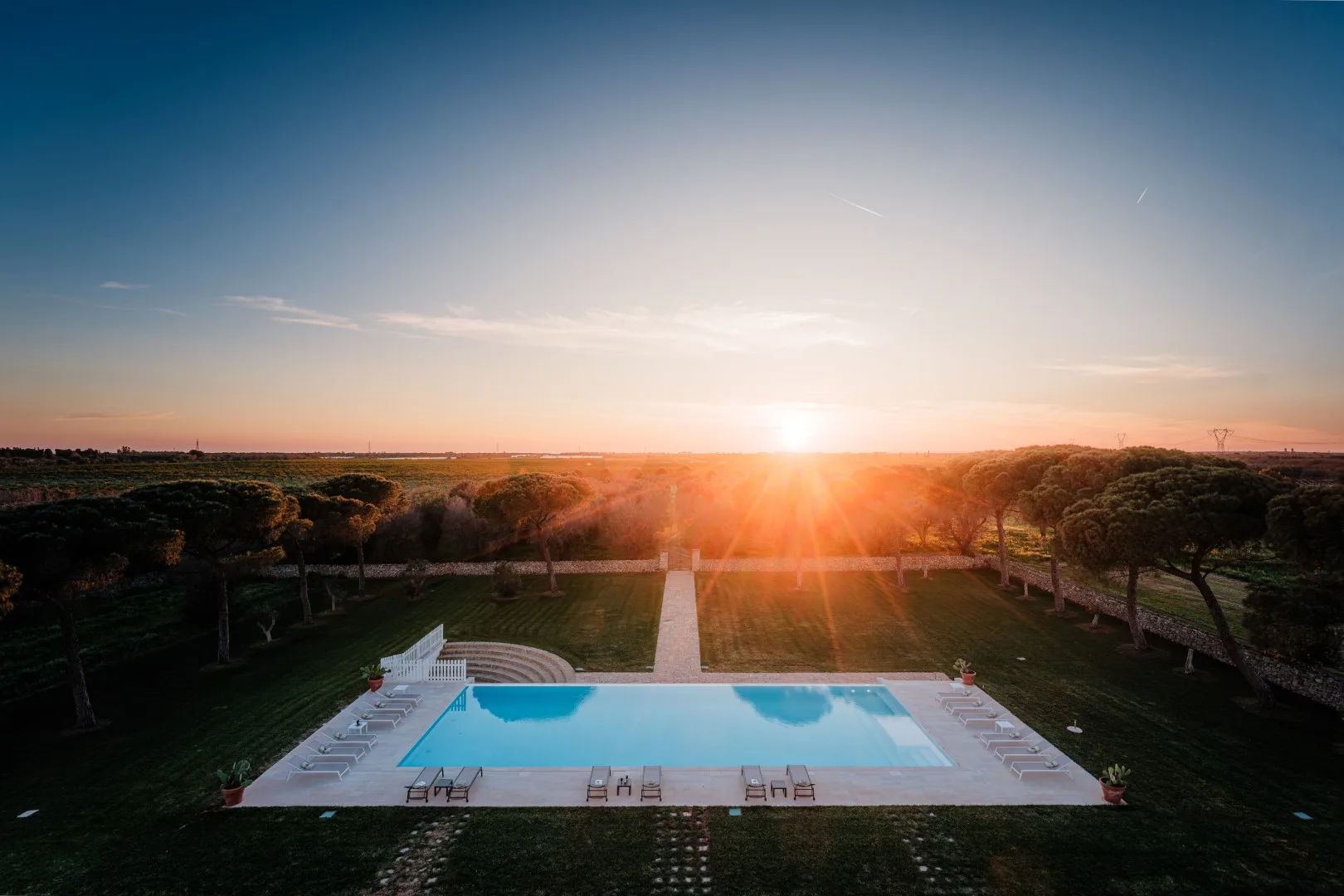 Una piscina rectangular y una terraza pavimentada están rodeadas de árboles bajo un atardecer resplandeciente.