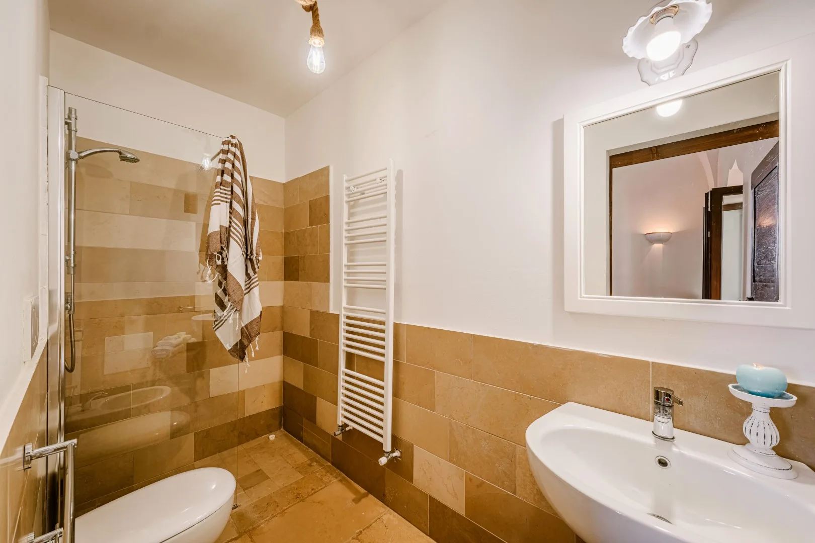 Un baño alicatado cuenta con una ducha de vidrio, un toallero calefactable y un lavabo blanco bajo un espejo.