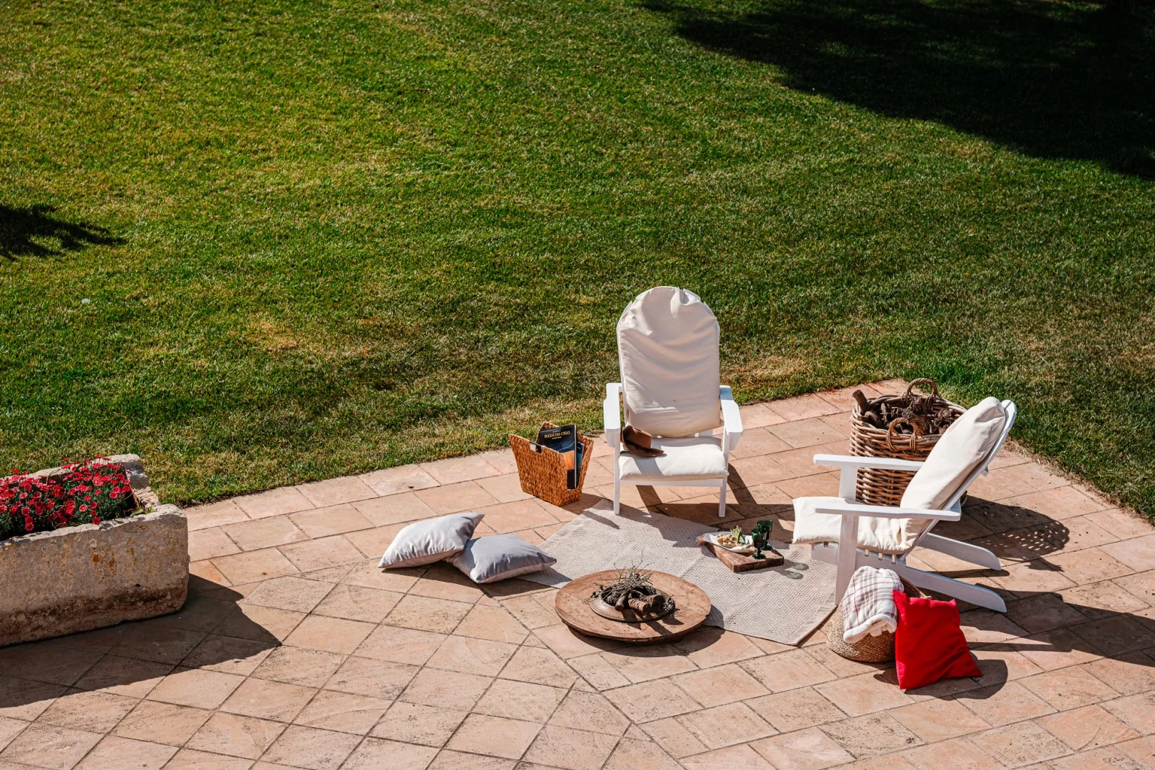 Un patio de baldosas tiene dos sillas blancas, cojines, un brasero y una jardinera junto a un césped.