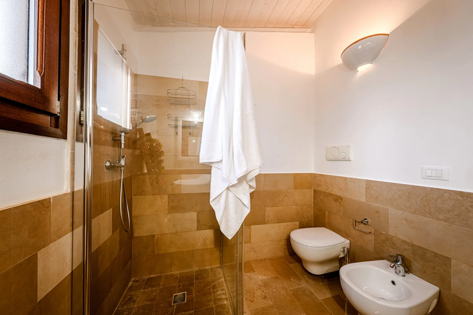 Un baño revestido de azulejos con ducha de vidrio, inodoro suspendido y lavabo blanco.
