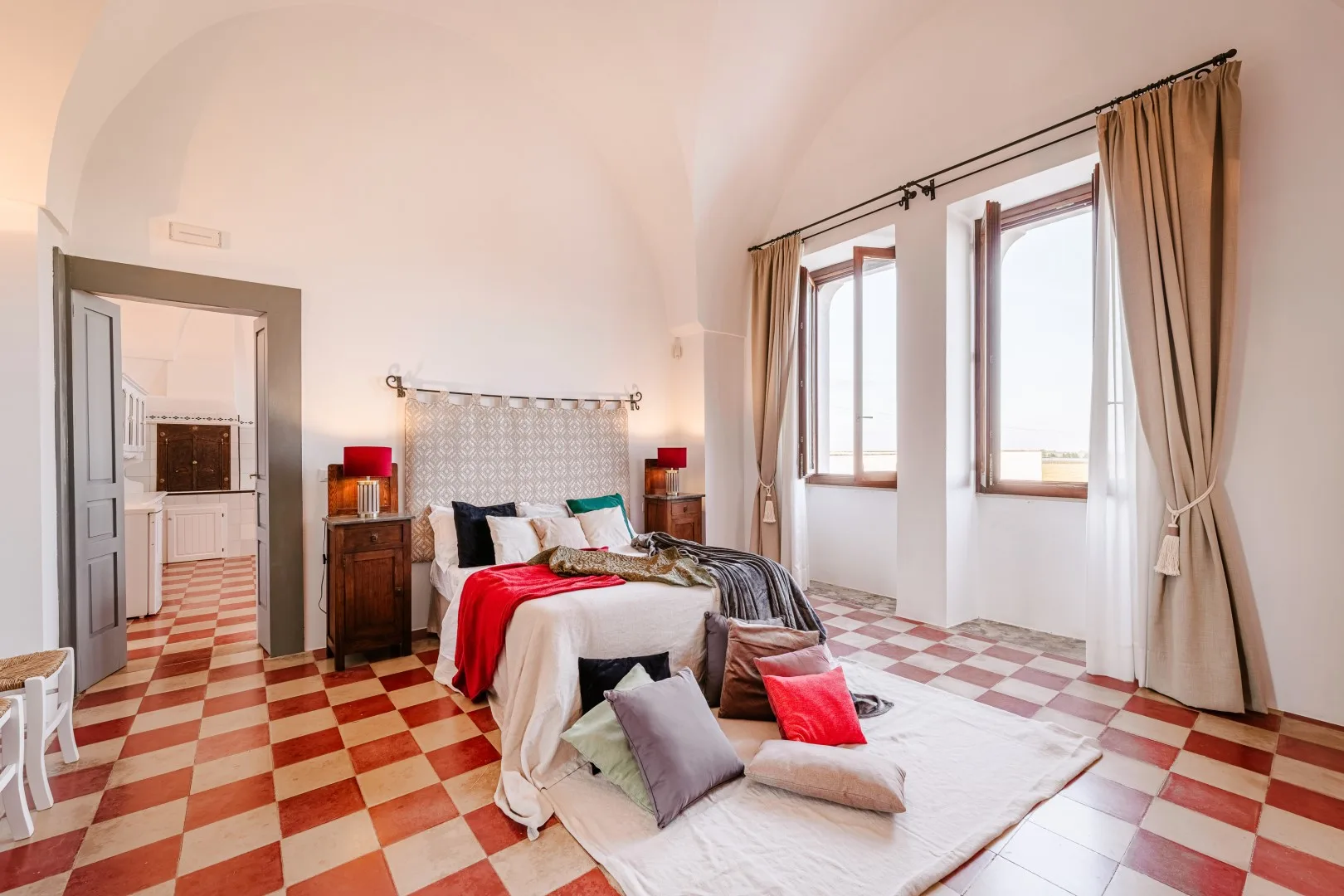 Dormitorio con cama blanca, ropa de cama roja y gris, lámparas de noche y ventanas altas con cortinas.