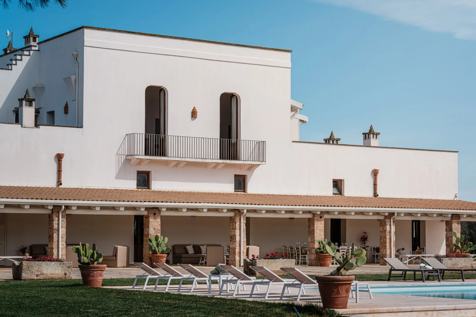 Un edificio blanco da a un patio junto a la piscina con tumbonas, plantas en macetas y una zona cubierta para sentarse.
