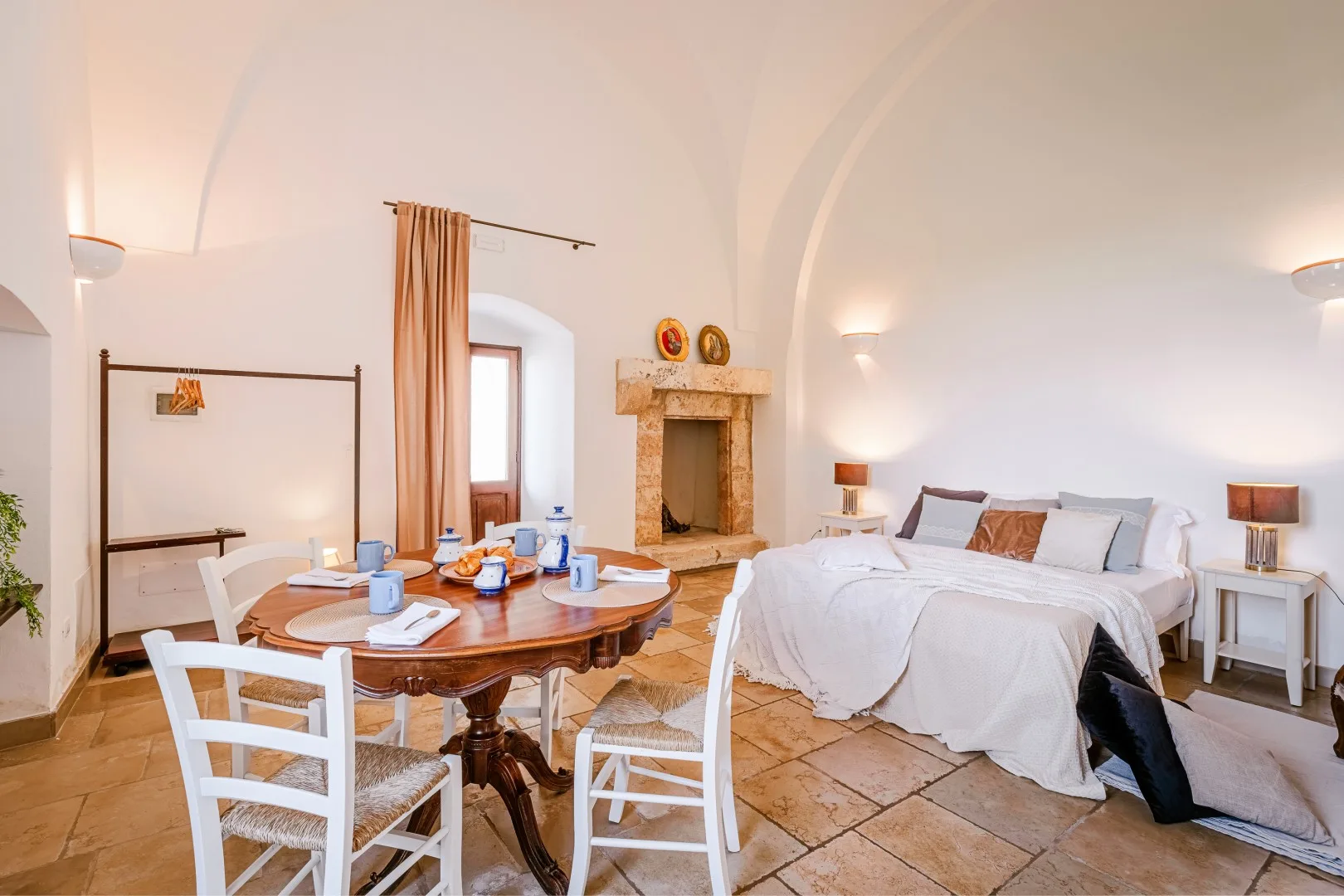 Un dormitorio luminoso con una cama blanca, una chimenea de piedra y una mesa de comedor de madera con sillas blancas.