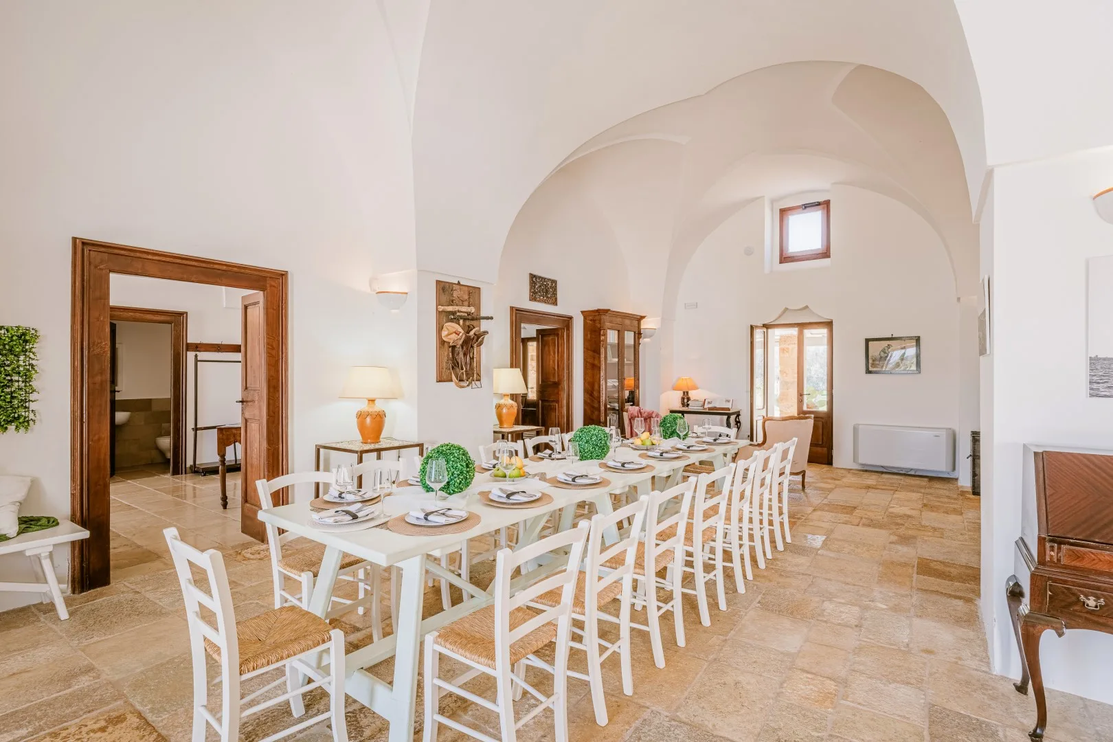 Una larga mesa de comedor con sillas blancas está preparada bajo los altos techos abovedados de una estancia luminosa.