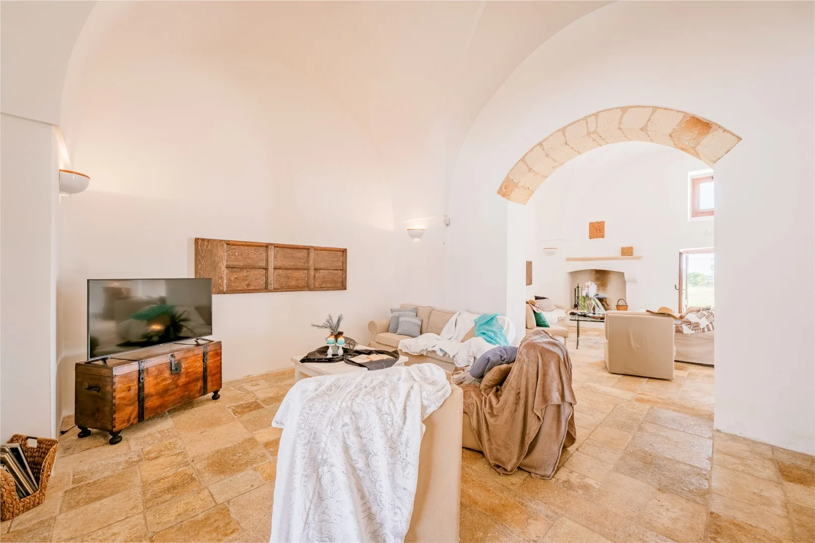 Una sala luminosa con televisor, sofás claros, un mueble bajo de madera y una abertura arqueada.