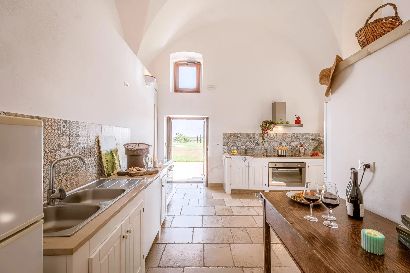 Una cocina luminosa tiene techo abovedado, encimeras de azulejos, una isla central y una puerta abierta al exterior.