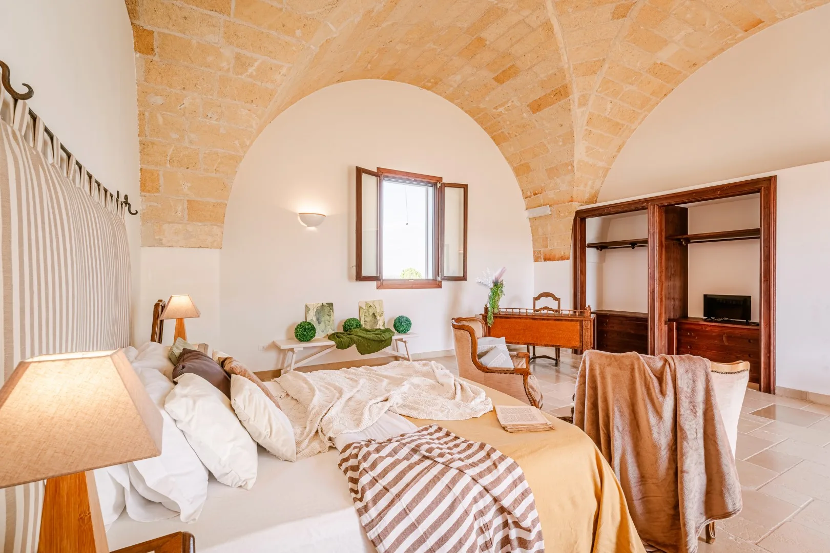 Dormitorio con una cama grande bajo un techo abovedado de ladrillo, una ventana y estanterías de madera empotradas.