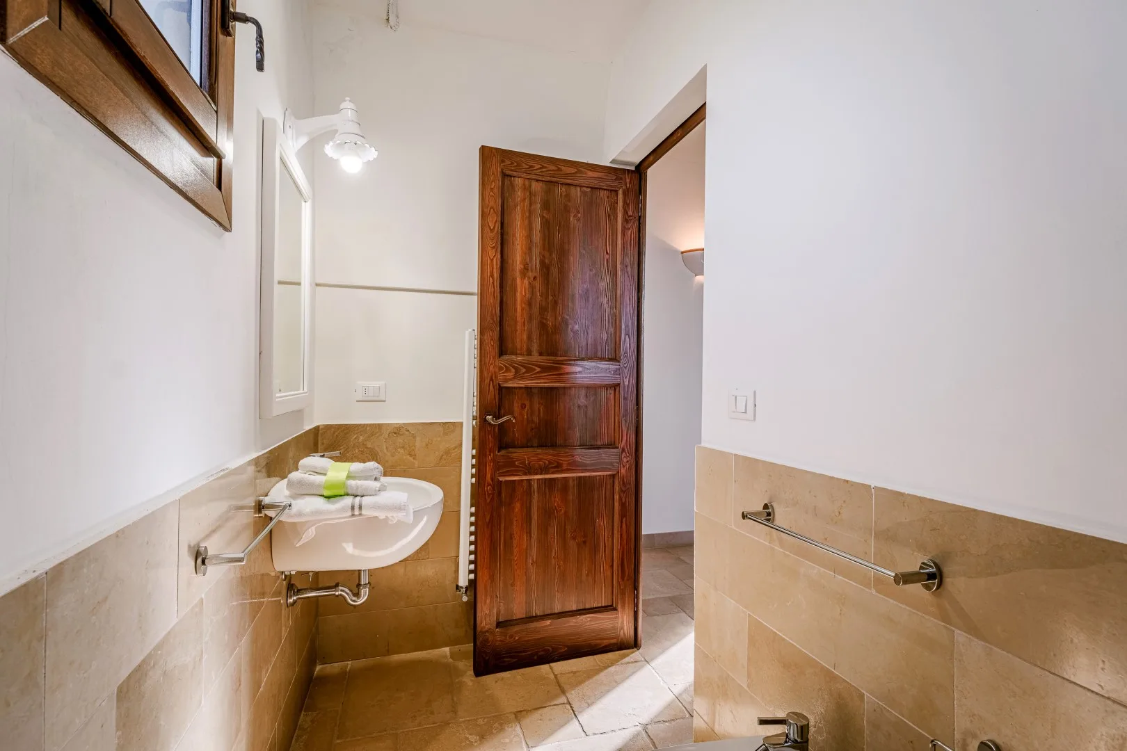 Un baño con azulejos tiene un lavabo blanco, un grifo de pared y una puerta de madera abierta.
