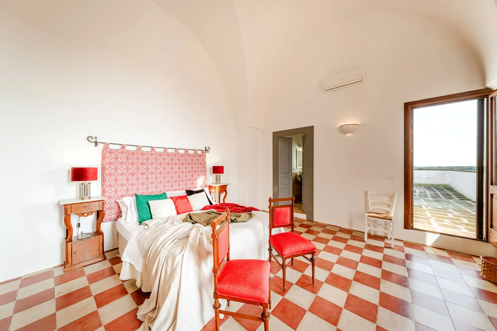 Un dormitorio luminoso tiene una cama blanca, sillas rojas, cortinas estampadas y suelo de baldosas rojas y crema.