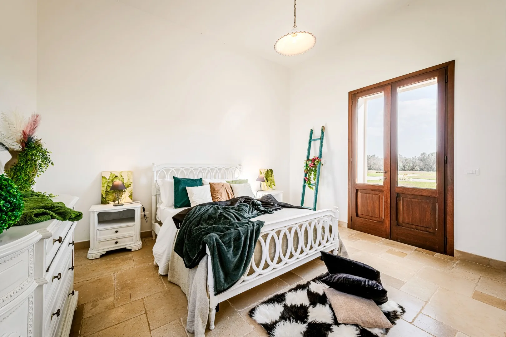Un dormitorio luminoso con una cama de metal blanco, ropa de cama verde oscuro y puertas francesas de madera que dan a un balcón.
