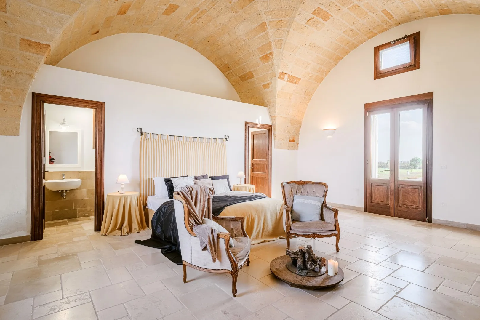 Un dormitorio luminoso con techo abovedado de piedra, cama doble, dos sillones y altas puertas acristaladas.