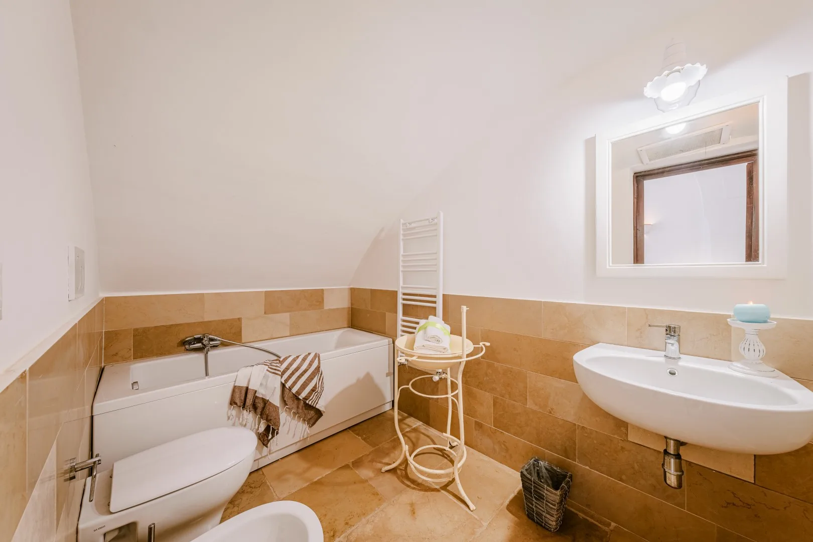 Un baño con bañera, lavabo blanco y azulejos color beige en la parte baja de las paredes y el suelo.
