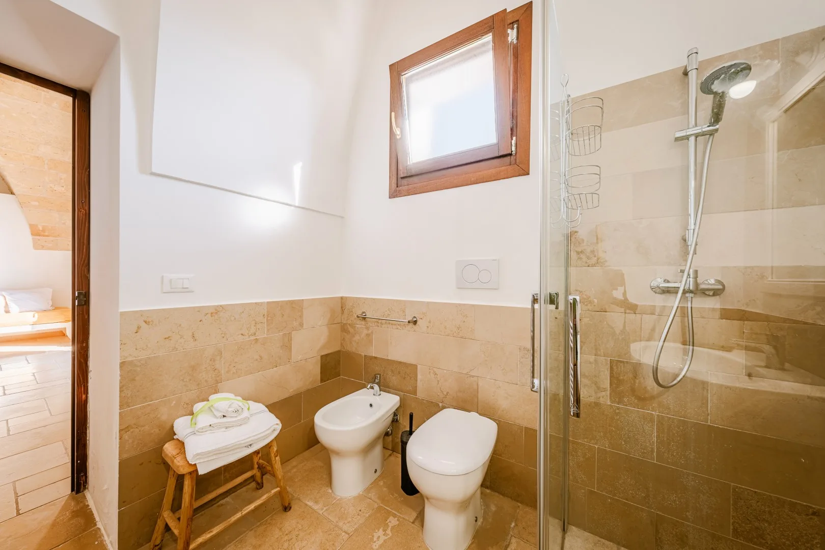 Un baño alicatado tiene una ducha con mampara de vidrio, un bidé, un inodoro y toallas dobladas sobre un taburete de madera.