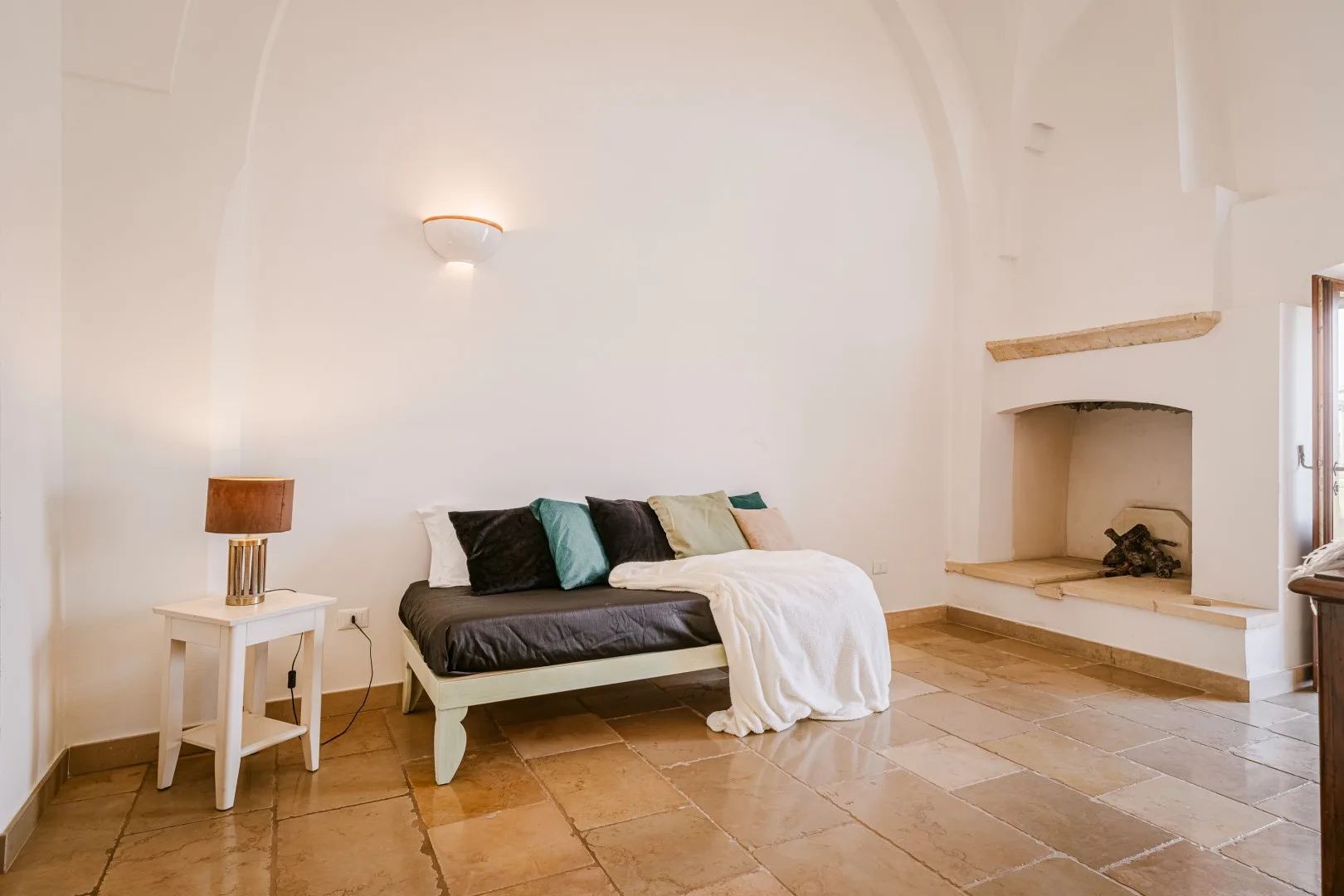 Una cama con varias almohadas y una manta blanca está junto a una mesita en una amplia habitación de suelo de baldosas.