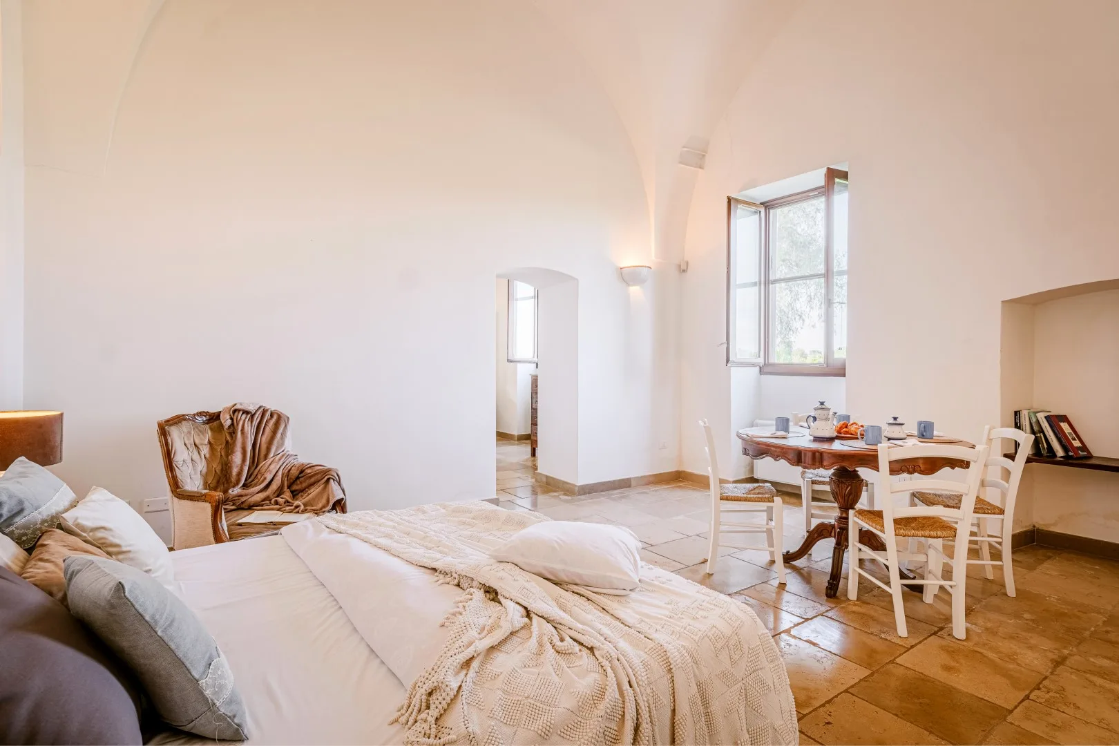Un dormitorio luminoso con una cama grande, un sillón y una pequeña mesa de comedor junto a ventanas altas.