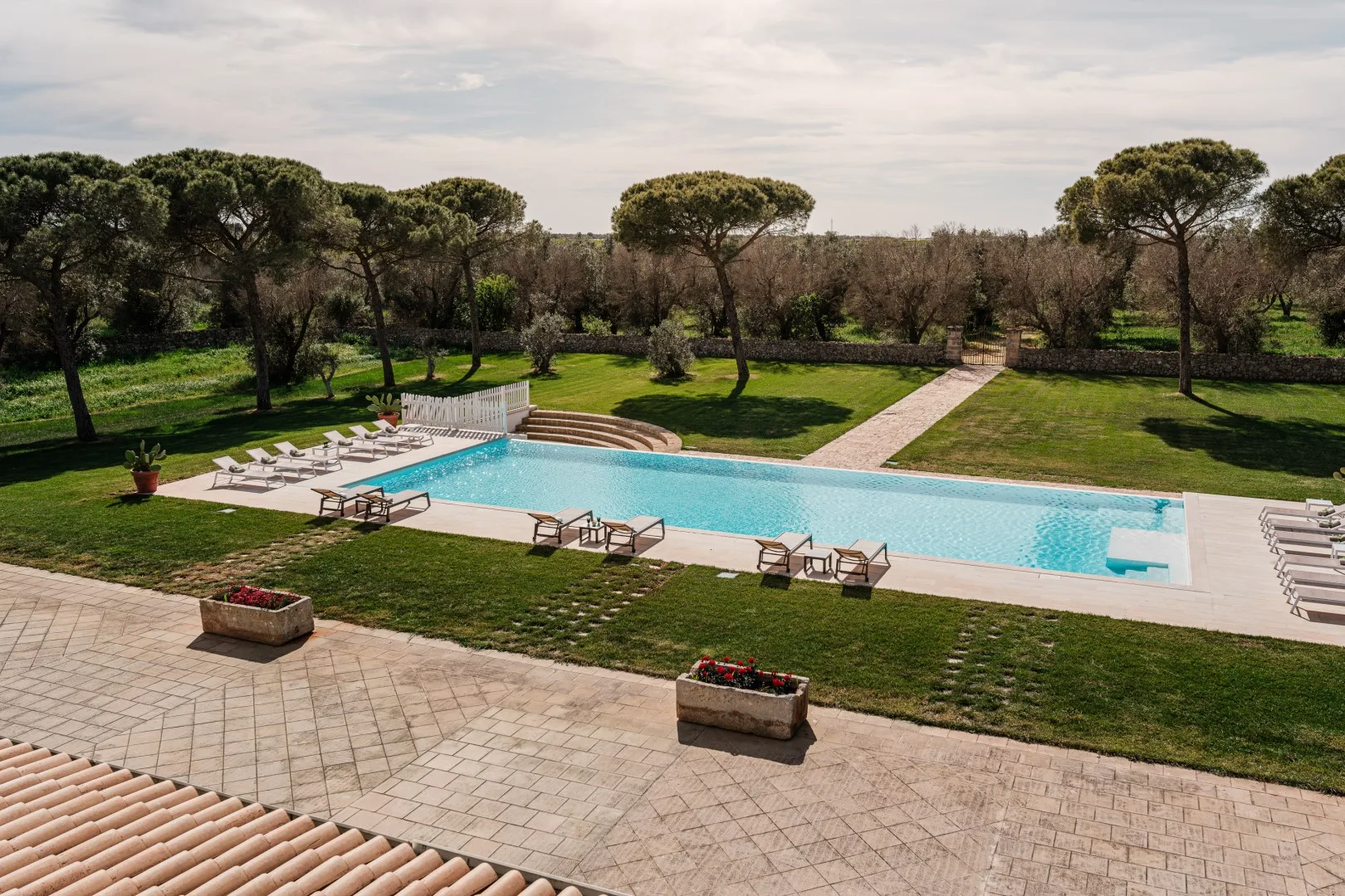 Una piscina rectangular al aire libre está rodeada de tumbonas, terrazas pavimentadas y jardines.