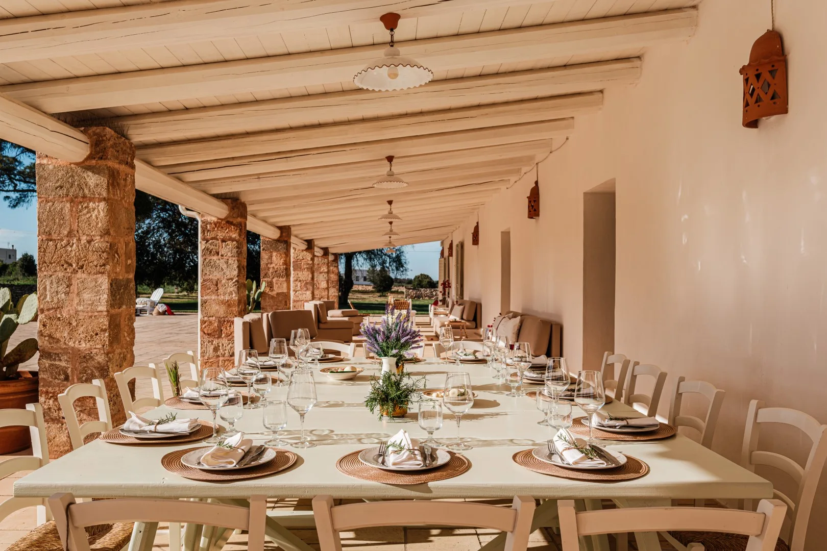 Una larga mesa está preparada con platos y copas bajo una veranda cubierta con columnas de piedra.