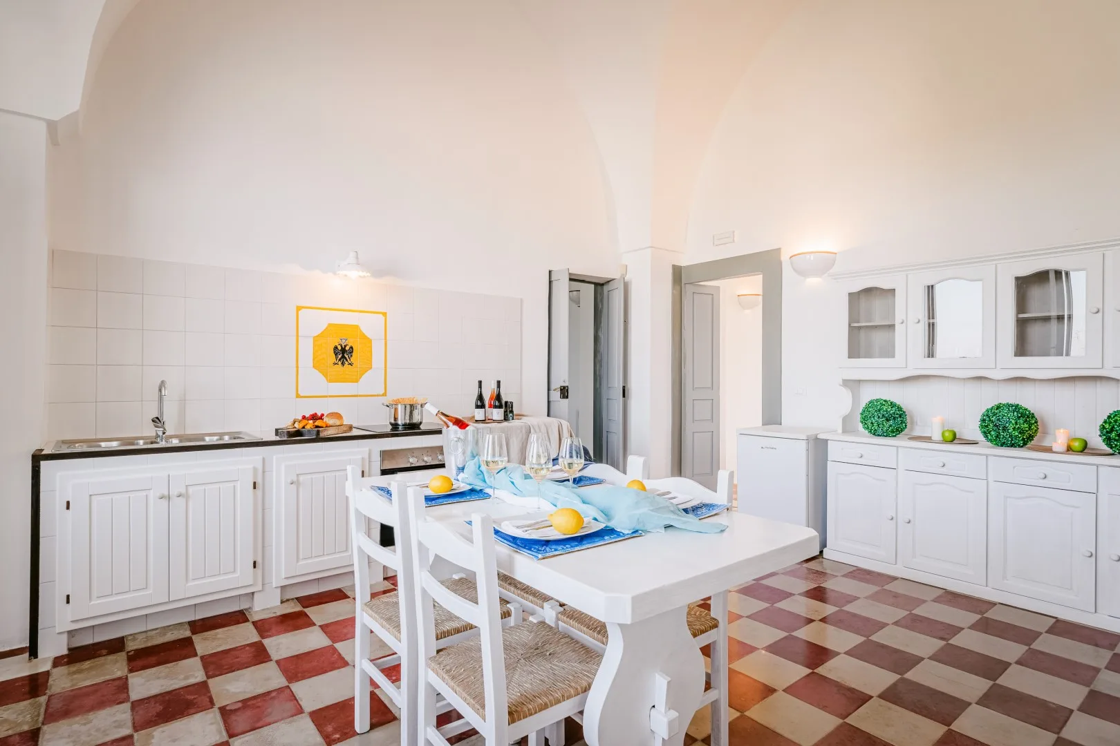Una cocina luminosa con armarios blancos, suelo de baldosas y una mesa puesta con vajilla azul.