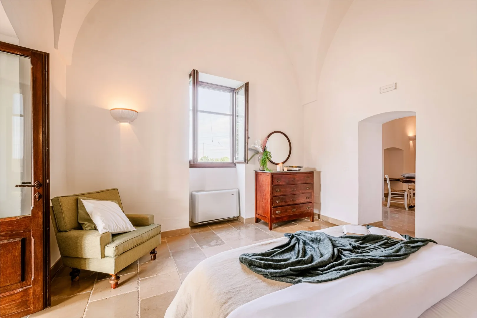 Un dormitorio con una cama, un sillón verde oliva, una cómoda de madera y una ventana abierta.