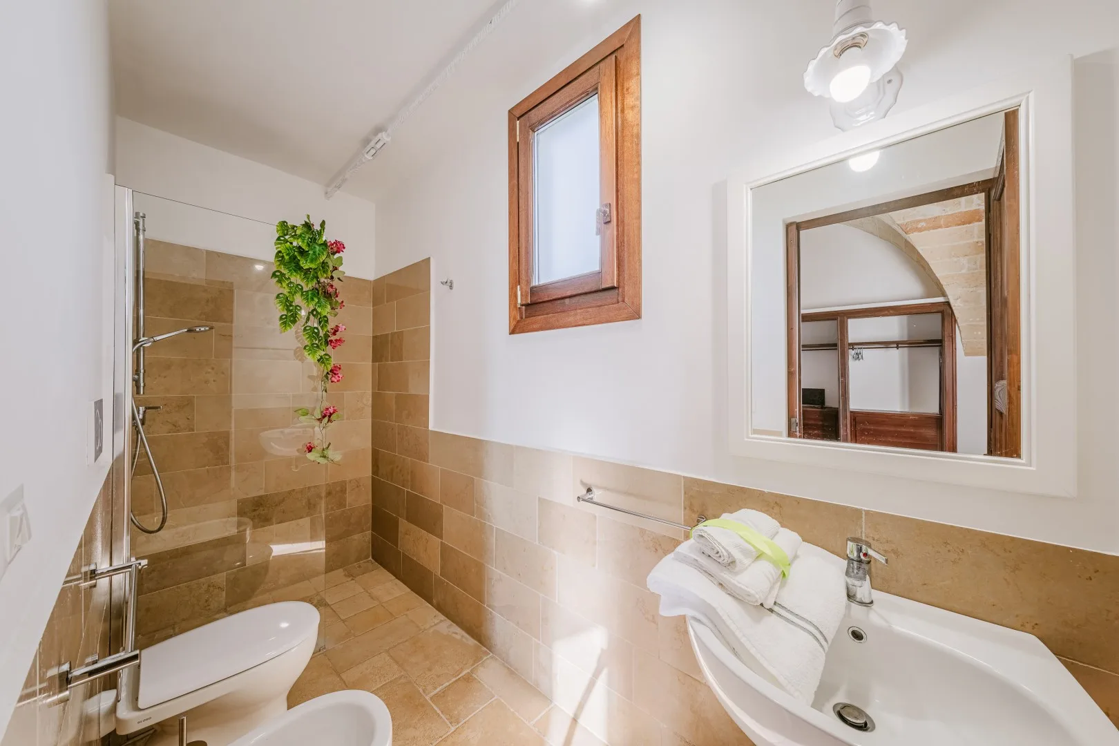 Un baño de azulejos cuenta con ducha acristalada, espejo, ventana pequeña y lavabo con toallas dobladas.