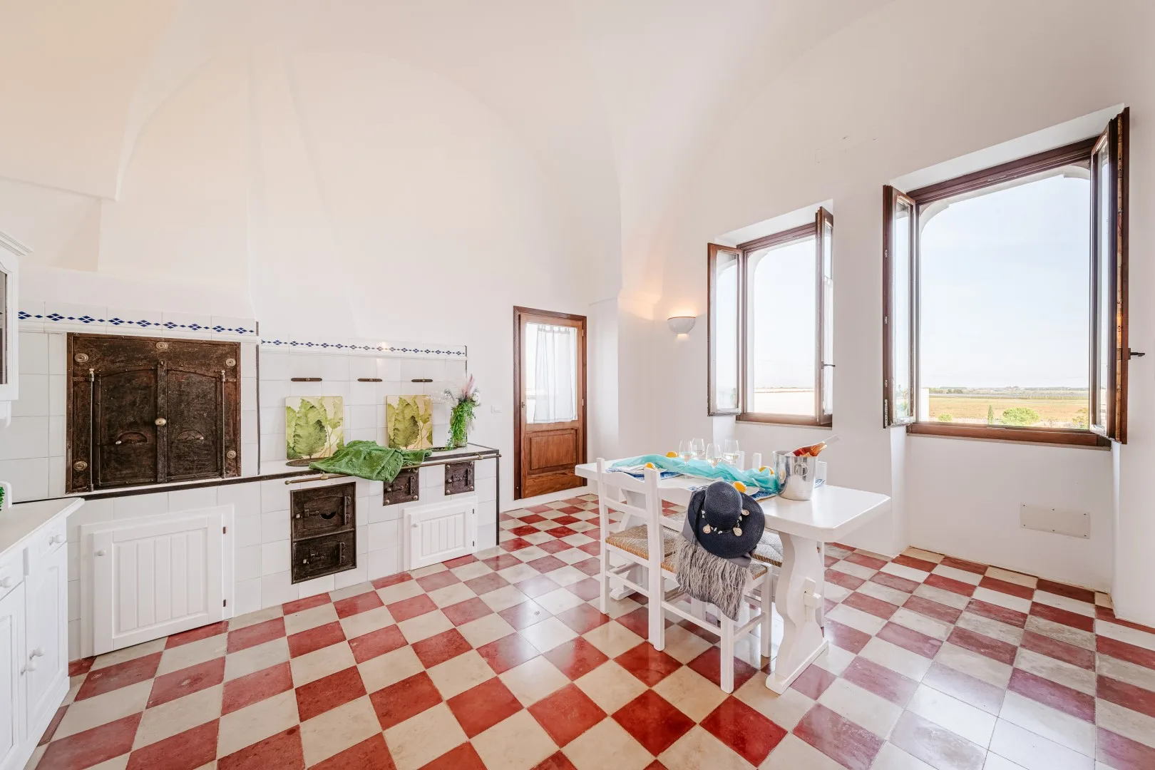 Una cocina luminosa tiene armarios blancos, suelo de baldosas a cuadros y una mesa junto a ventanas abiertas.