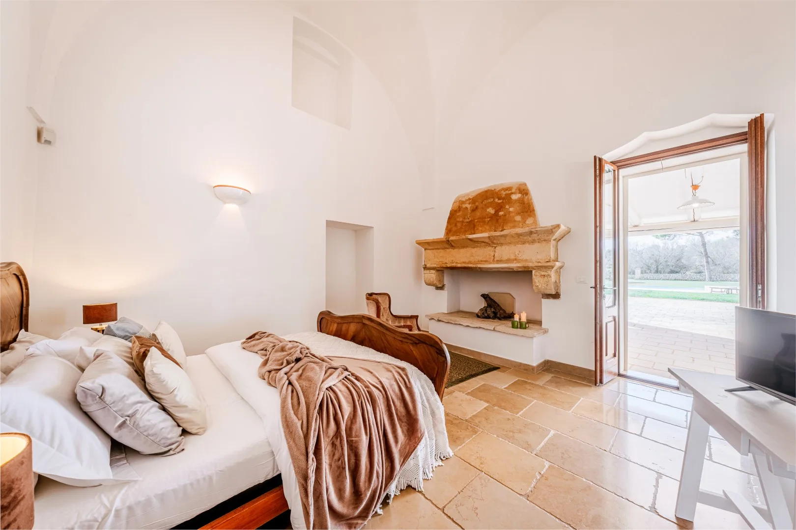 Un dormitorio luminoso tiene una cama grande, una chimenea de piedra y puertas abiertas que dan a un patio.