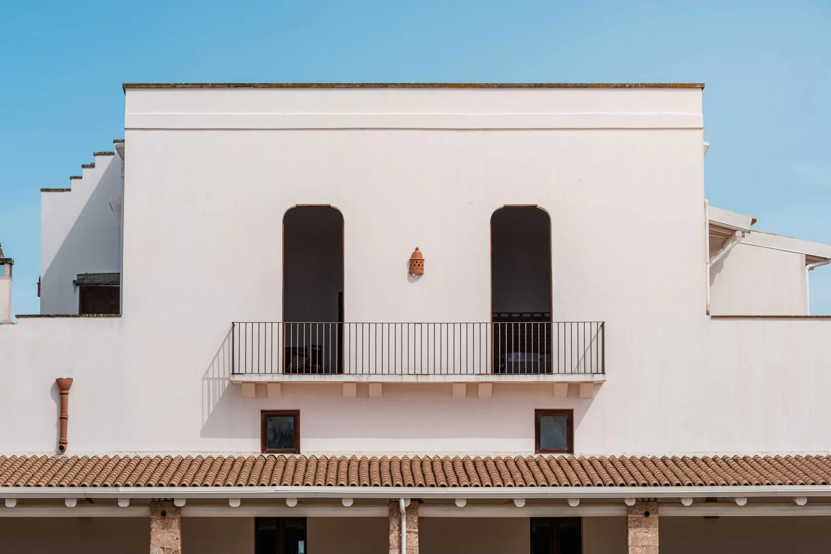 Un edificio blanco de dos plantas tiene un balcón estrecho con barandilla negra y dos puertas arqueadas.
