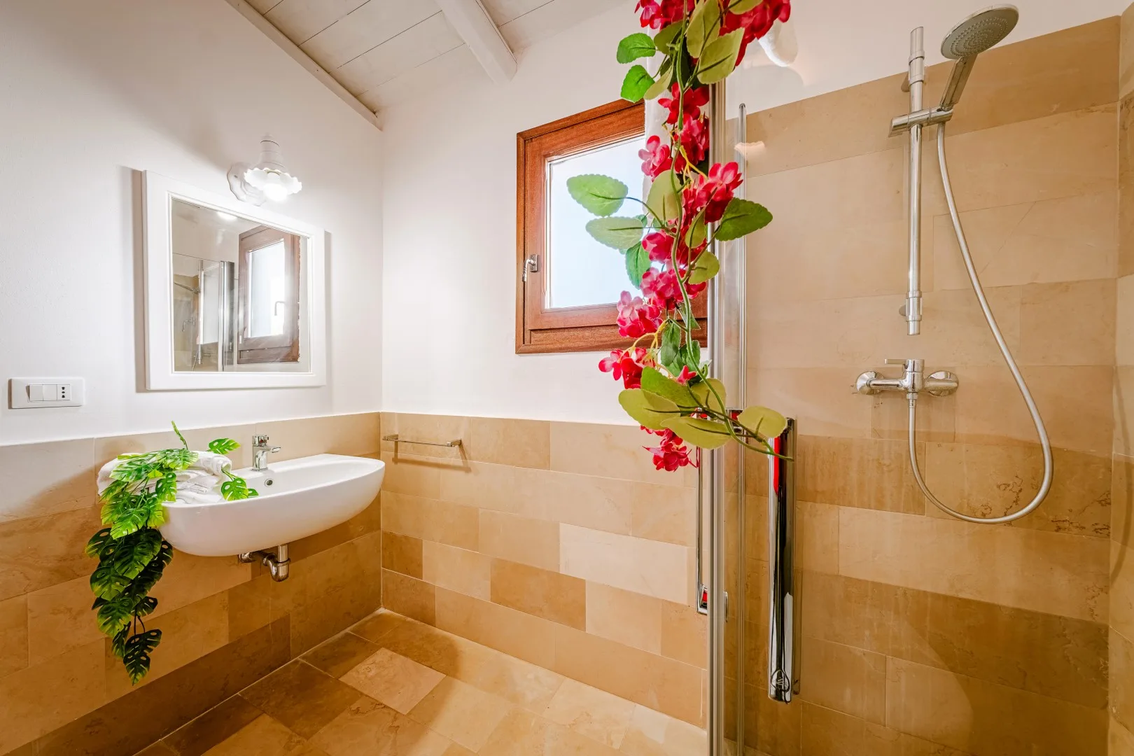 Un baño alicatado cuenta con un lavabo suspendido, una ducha con mampara de vidrio y una ventana con flores rojas.