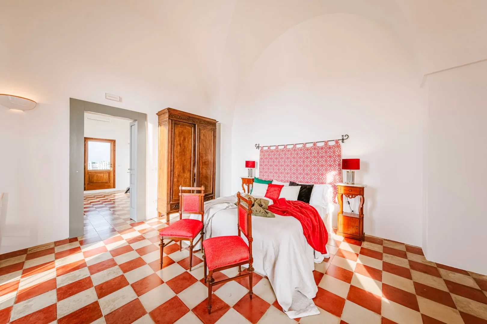 Un dormitorio luminoso tiene una cama blanca con detalles rojos, muebles de madera y un suelo ajedrezado rojo y blanco.
