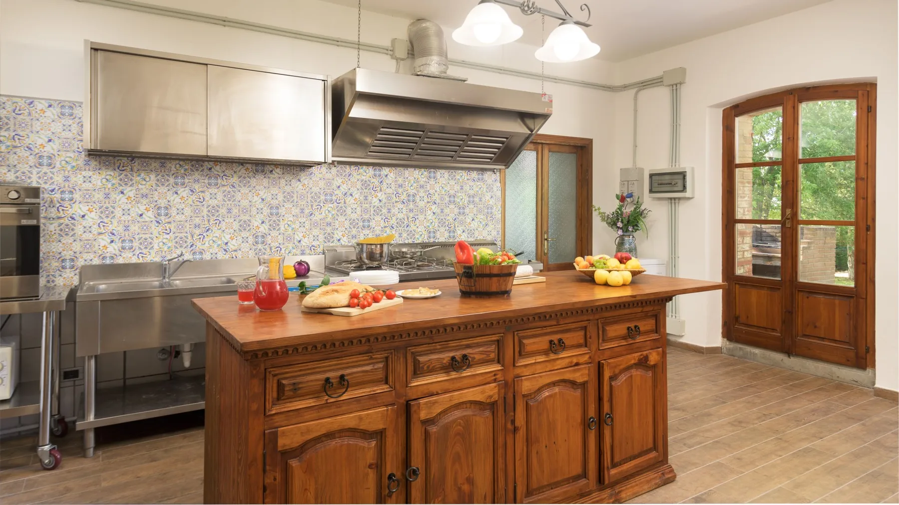 Una cocina con azulejos tiene electrodomésticos de acero inoxidable, una isla de madera con comida y puertas acristaladas.