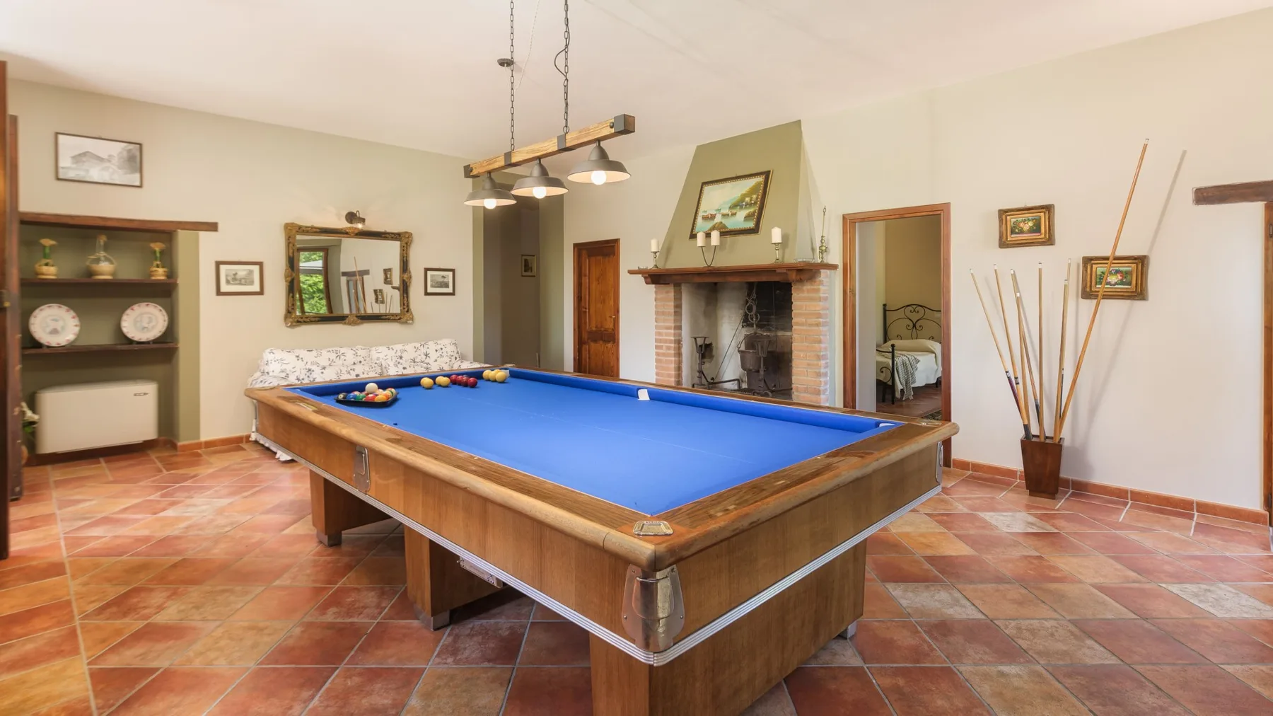 Una mesa de billar con paño azul ocupa una sala con suelo de baldosas, chimenea, estantes y cuadros.